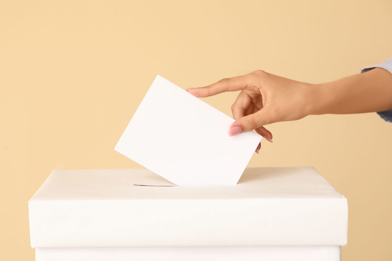 Das Foto zeigt eine Hand, die einen Wahlzettel in eine Urne schmeißt.